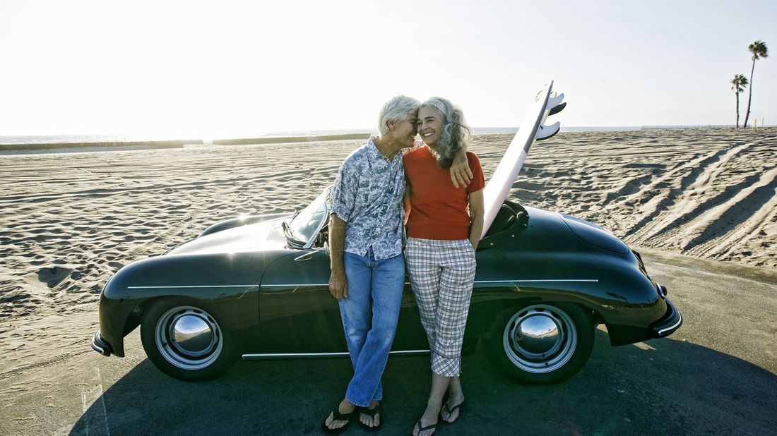 Retired couple posing in front of a car at the beach.