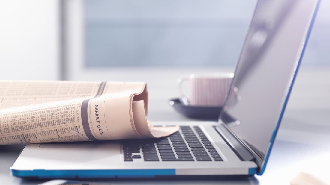 An open laptop with a newspaper open to the markets section