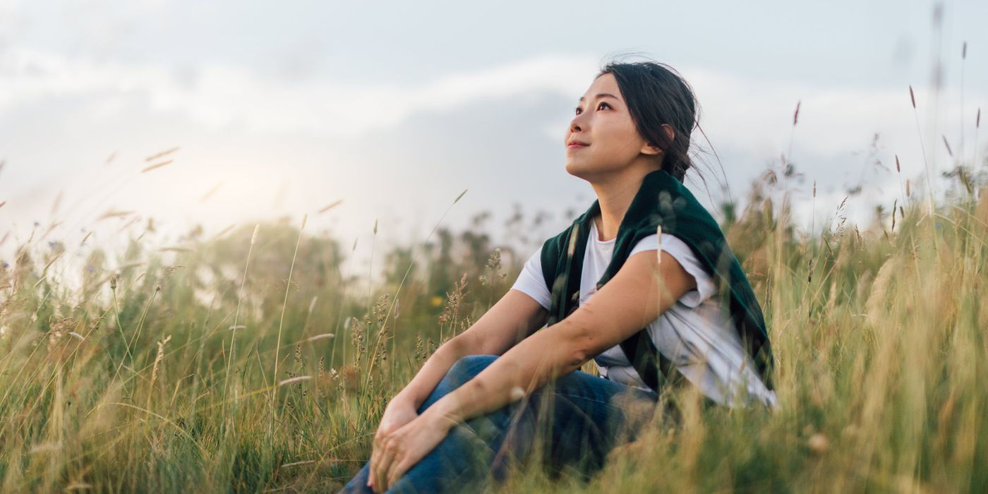 Woman looking up to the sky while sitting in meadow, enjoying a relaxing afternoon in natural parkland against sunlight.