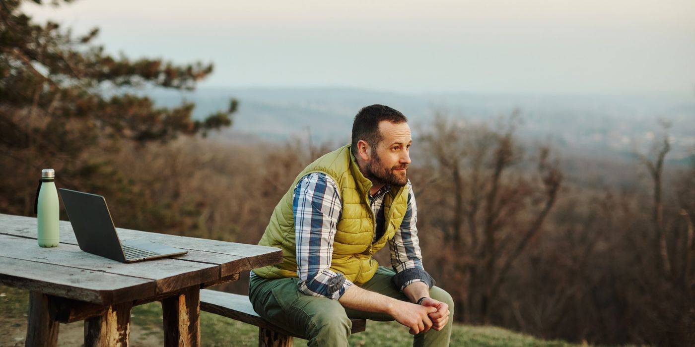 A photo of a man sitting on a wooden bench on a high hill overlooking the city. Taking a break from working on his laptop, he rests by looking at the natural beauty around him. 