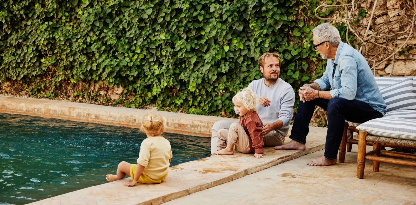 Father and son in discussion while relaxing with family on poolside terrace of tropical villa during multigenerational family vacation 