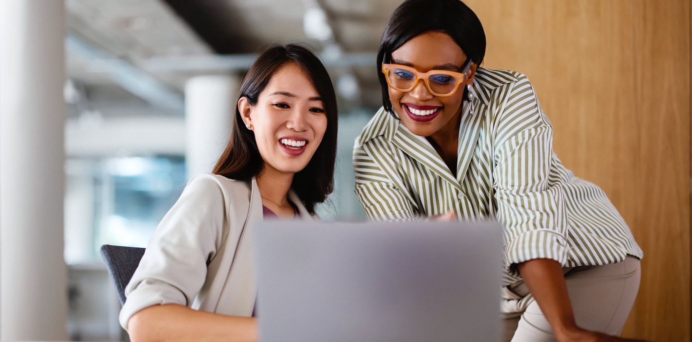 Two professional women discussing work on a laptop, demonstrating teamwork and collaboration in a modern office setting.  