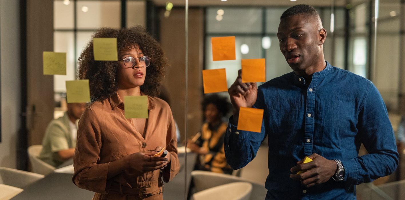 Smart young workers of IT company discussing plans in meeting room. Putting stickers with ideas on glass wall during brainstorming.