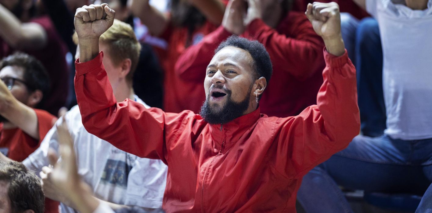 Adult male soccer fan passionately cheers and raises his arms while standing in a large crowd of supporters