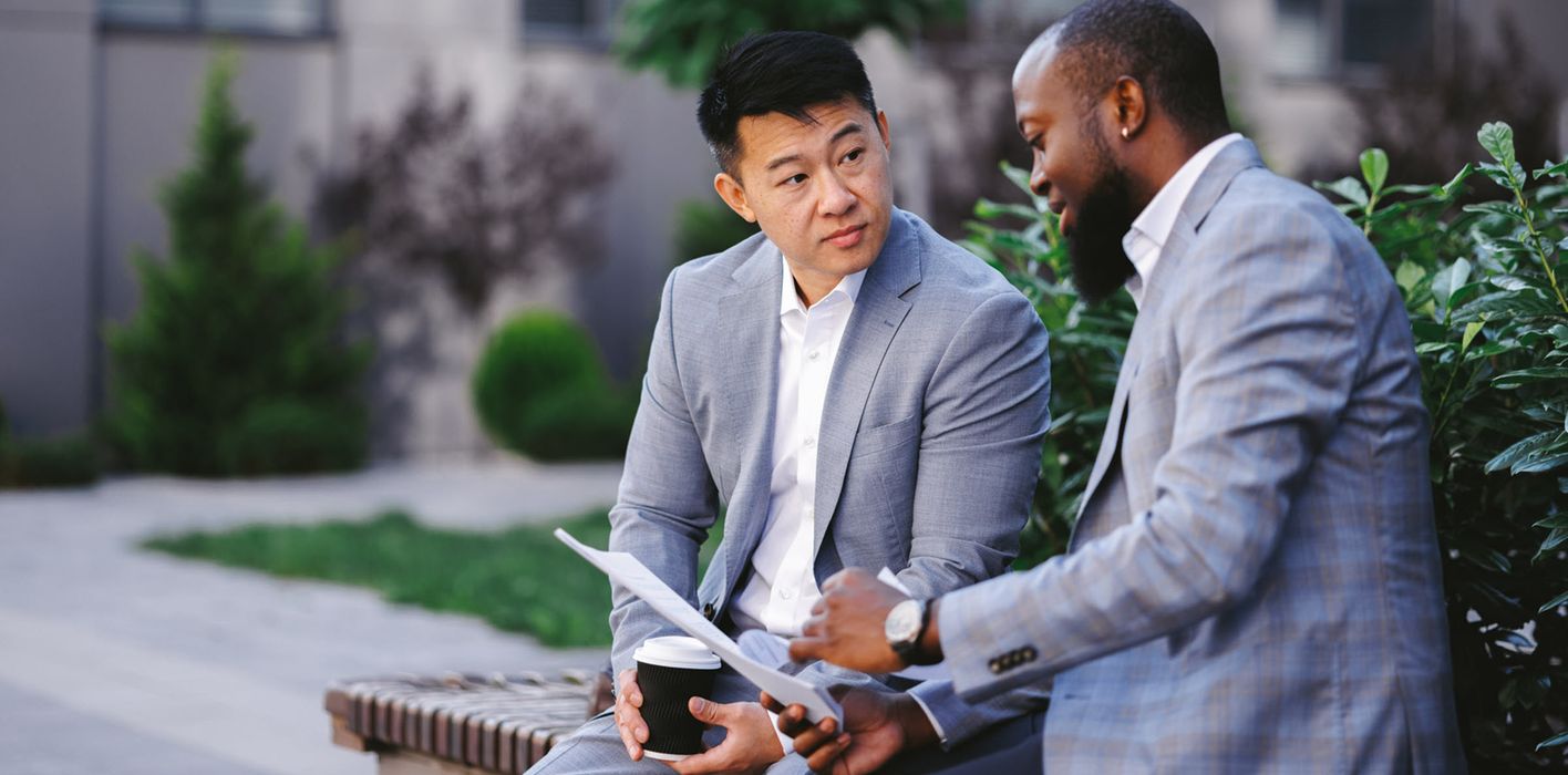 Side view of two colleagues outside office, reading information on a document and drinking coffee