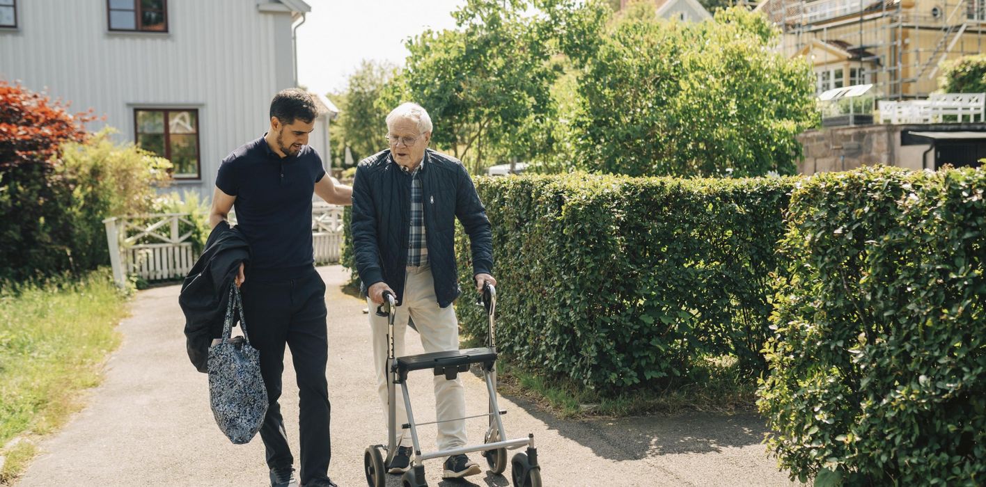 Male caregiver walking with senior man using rollator in park at sunny day