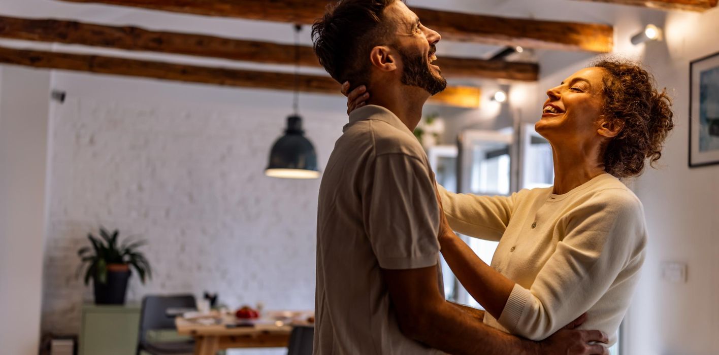 joyful couple shares a warm hug and big smile in a bright, cozy living space, conveying love, connection, and positive energy in a home environment.