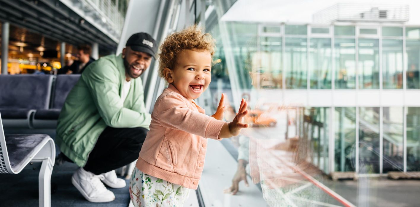 A joyful toddler girl presses her hands against the airport glass as her laughing father crouches behind her