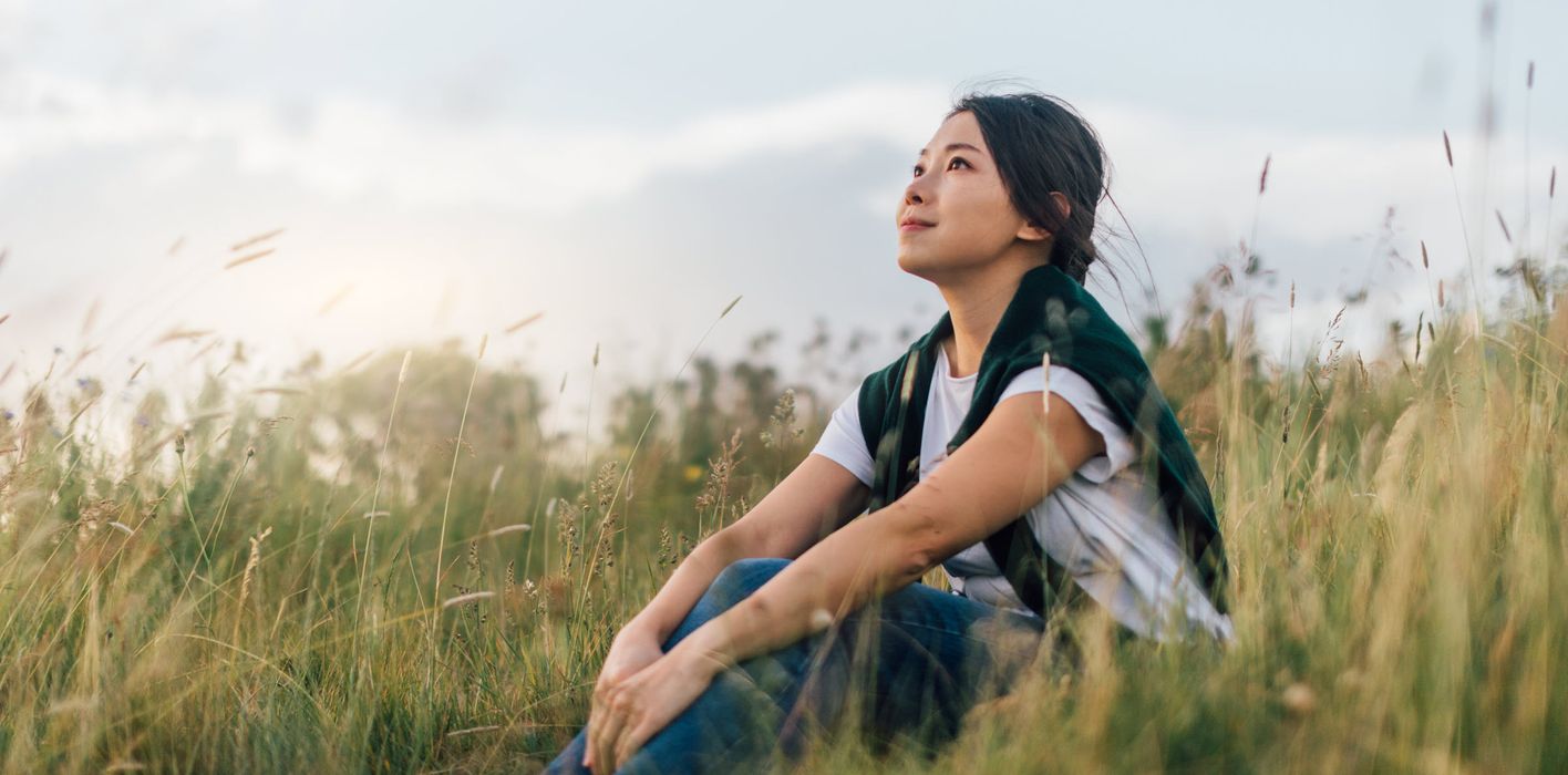 Woman looking up to the sky while sitting in meadow, enjoying a relaxing afternoon in natural parkland against sunlight.