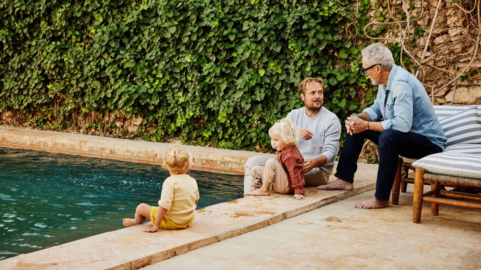 Father and son in discussion while relaxing with family on poolside terrace of tropical villa during multigenerational family vacation 