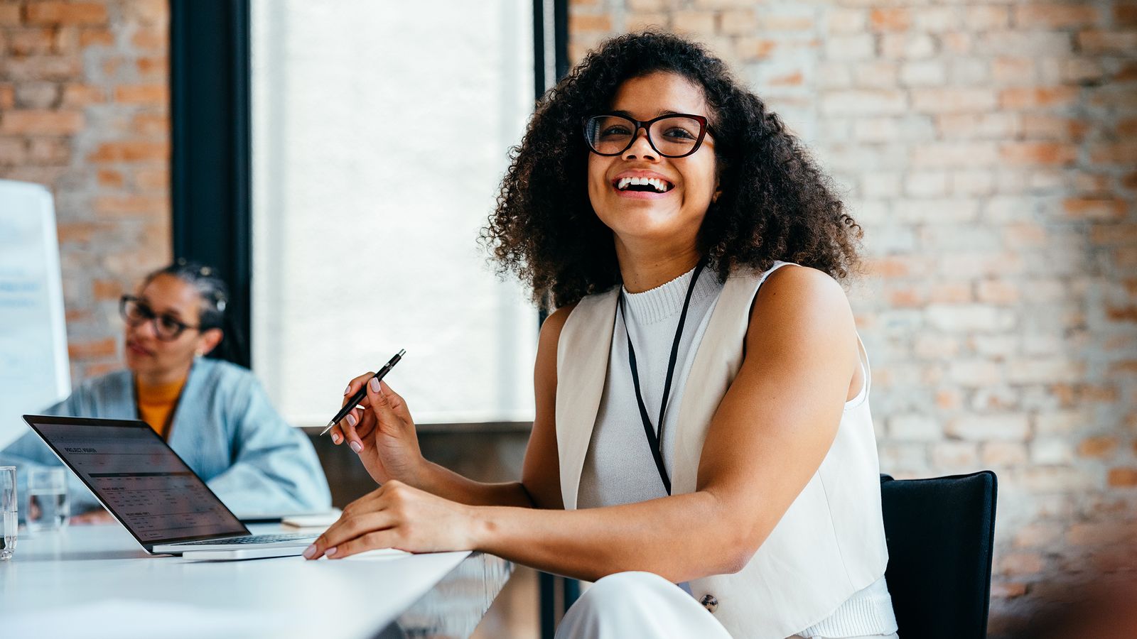 Confident young professional woman smiling in a modern office environment alongside colleague, working on a laptop and engaging in a business meeting.