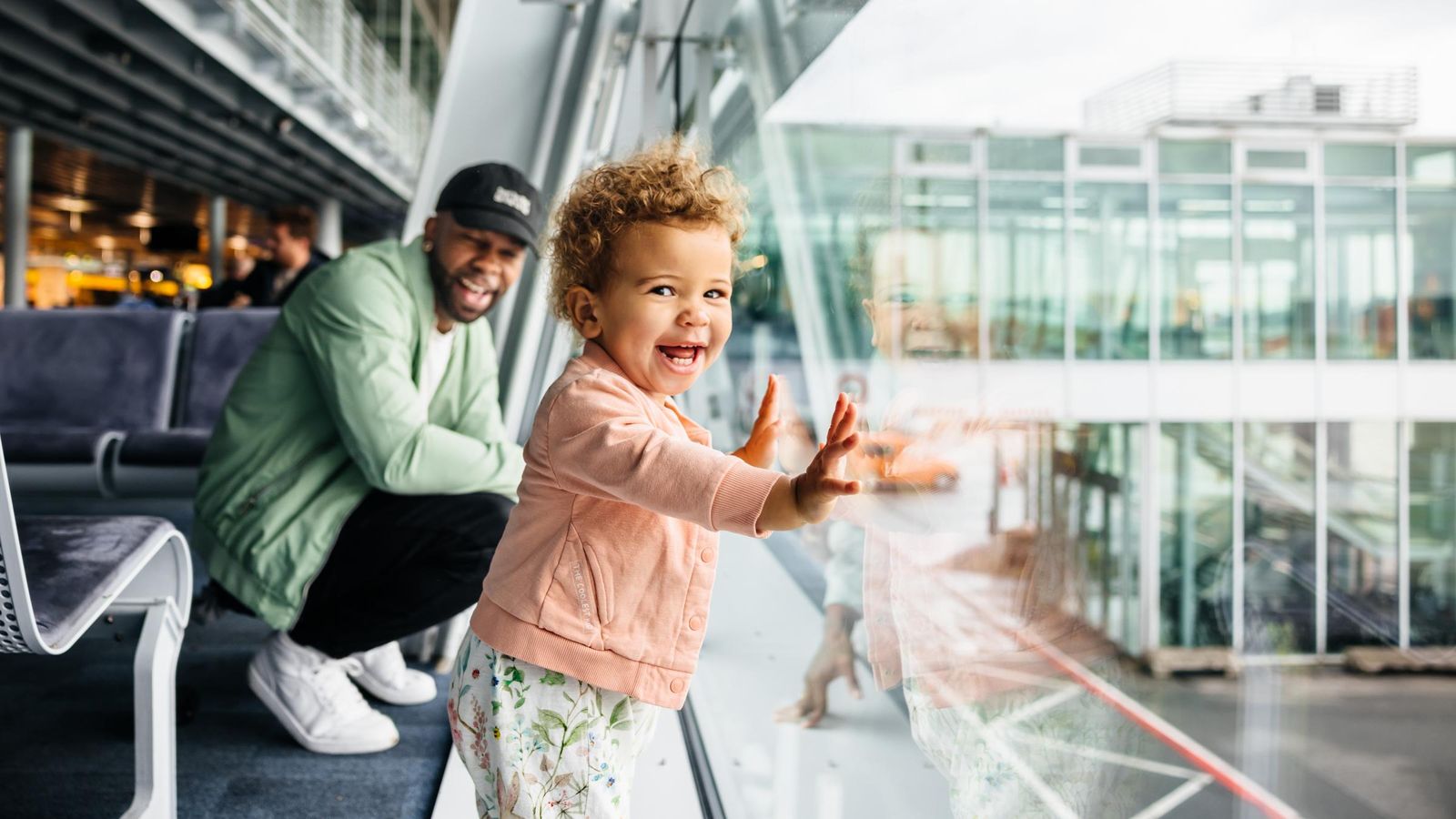 A joyful toddler girl presses her hands against the airport glass as her laughing father crouches behind her