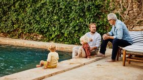 Father and son in discussion while relaxing with family on poolside terrace of tropical villa during multigenerational family vacation 