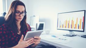 Woman looks at her tablet while a computer monitor displays graphs