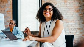 Confident young professional woman smiling in a modern office environment alongside colleague, working on a laptop and engaging in a business meeting.