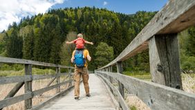 A father and young son walk on a bridge in a scenic outdoor setting.