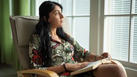 A woman reading a book in a rocking chair, looking through window with sunset light.