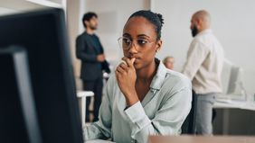 Contemplative young investment professional wearing eyeglasses sitting at desk while working on desktop PC in office reading Northwestern Mutual’s Weekly Market Commentary