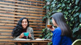 Cafe patio with lush foliage hosts two women enjoying coffee and pastries.