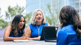Happy businesswomen in blue sat at table, laughing with laptop