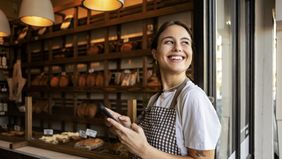 Young bakery owner standing proudly outside her shop using phone