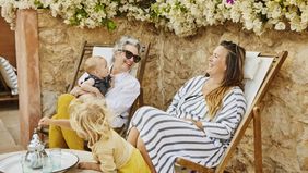 Wide shot of grandmother holding grandson in lap while laughing with daughter at tropical villa during multigenerational family vacation.