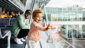 A joyful toddler girl presses her hands against the airport glass as her laughing father crouches behind her