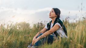 Woman looking up to the sky while sitting in meadow, enjoying a relaxing afternoon in natural parkland against sunlight.