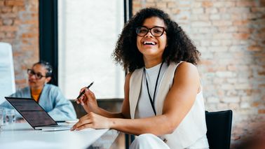 Confident young professional woman smiling in a modern office environment alongside colleague, working on a laptop and engaging in a business meeting.