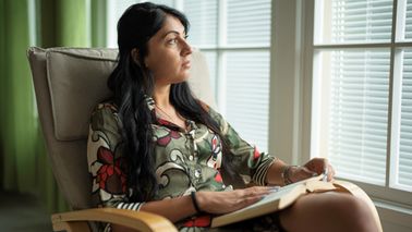 A woman reading a book in a rocking chair, looking through window with sunset light.