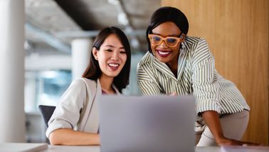 Two professional women discussing work on a laptop, demonstrating teamwork and collaboration in a modern office setting.  