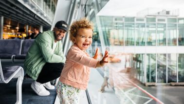 A joyful toddler girl presses her hands against the airport glass as her laughing father crouches behind her