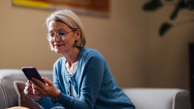 A woman wearing glasses smiles as she interacts with her smartphone, sitting casually in her home.