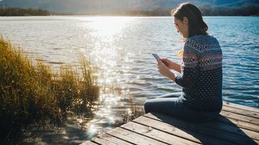 Woman sitting on a wooden platform at a lake at sunset, using smartphone