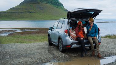 Woman and man sitting on car trunk drinking from travel tumbler during road trip on the background of Kirkjufell mountain in Iceland