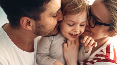 Close up shot of a cheerful couple hugging and kissing their smiling daughter in the middle.