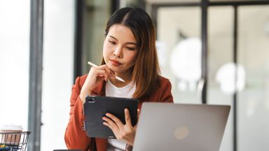 Young businesswoman looking at her digital tablet while sitting at a desk in stylish co-working space and working. Successful female entrepreneur and leadership. Technology in a modern lifestyle. Working remotely with flexibility. 