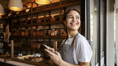 Young bakery owner standing proudly outside her shop using phone