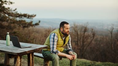 A photo of a man sitting on a wooden bench on a high hill overlooking the city. Taking a break from working on his laptop, he rests by looking at the natural beauty around him. 