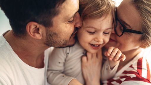 Close up shot of a cheerful couple hugging and kissing their smiling daughter in the middle.