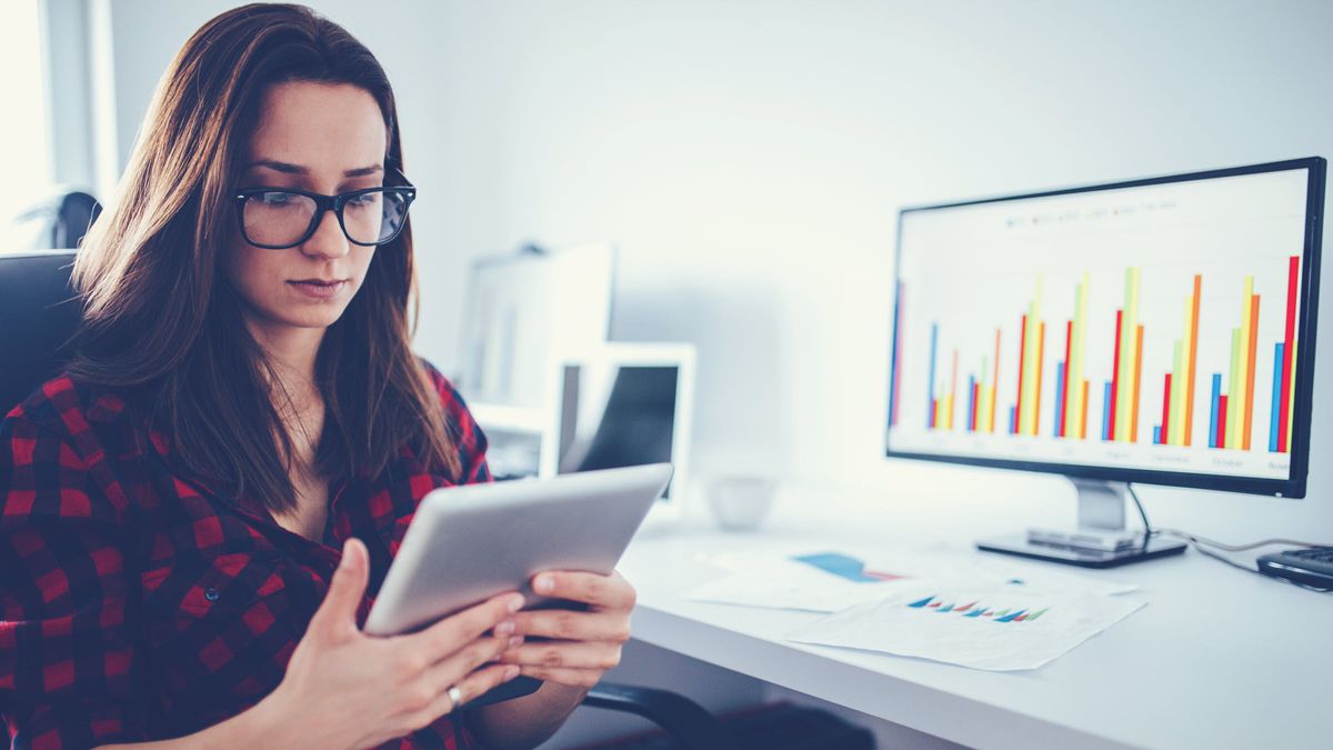 Woman looks at her tablet while a computer monitor displays graphs