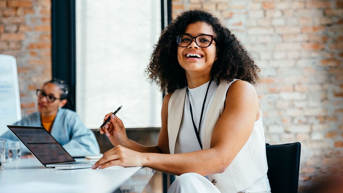 Confident young professional woman smiling in a modern office environment alongside colleague, working on a laptop and engaging in a business meeting.