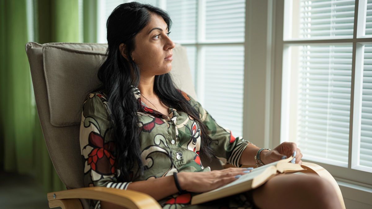 A woman reading a book in a rocking chair, looking through window with sunset light.