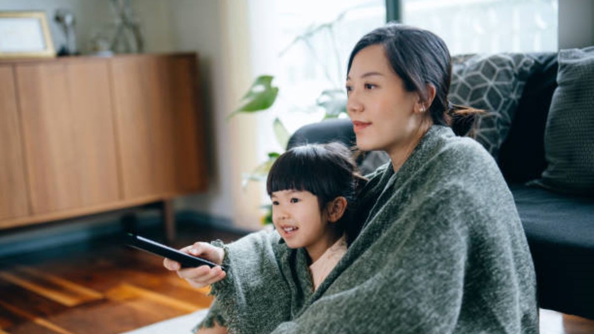 A mother and young daughter are wrapped in a blanket, relaxing at home and watching TV in the living room.