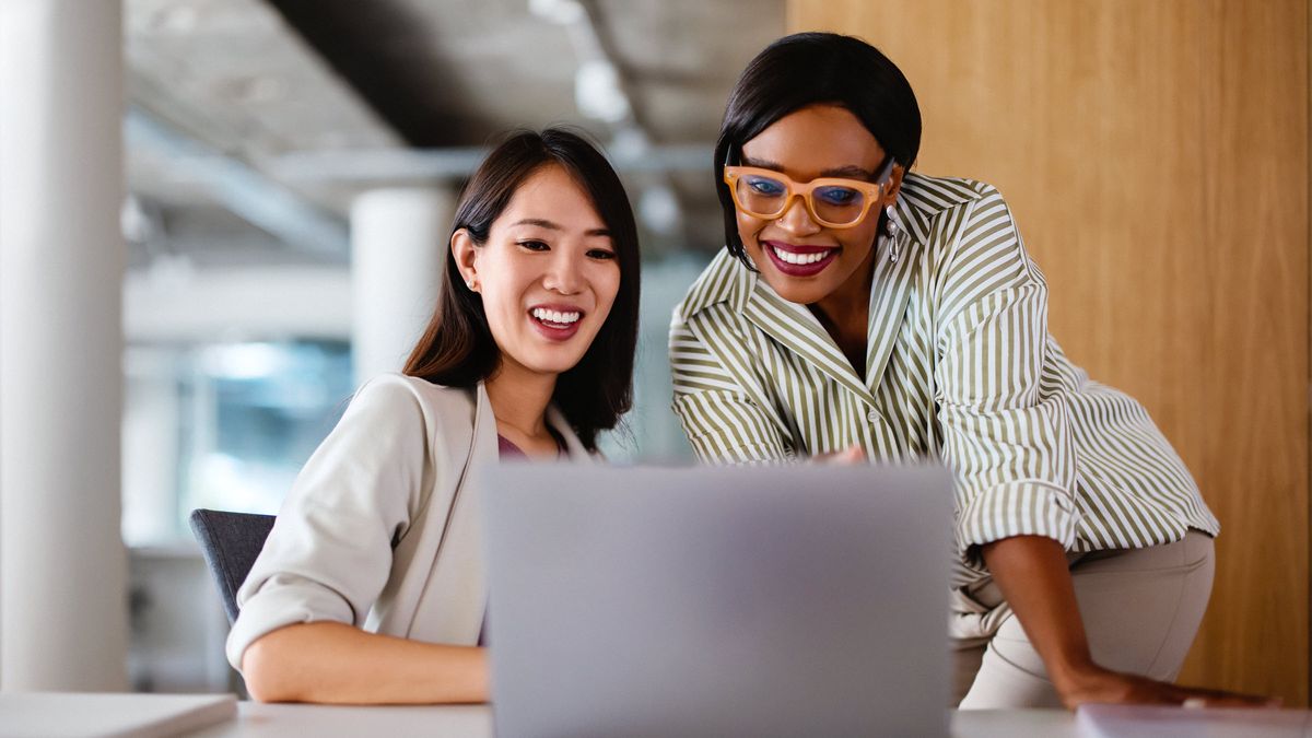 Two professional women discussing work on a laptop, demonstrating teamwork and collaboration in a modern office setting.  