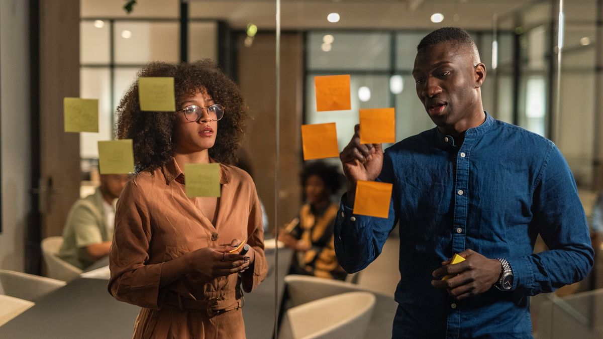 Smart young workers of IT company discussing plans in meeting room. Putting stickers with ideas on glass wall during brainstorming.