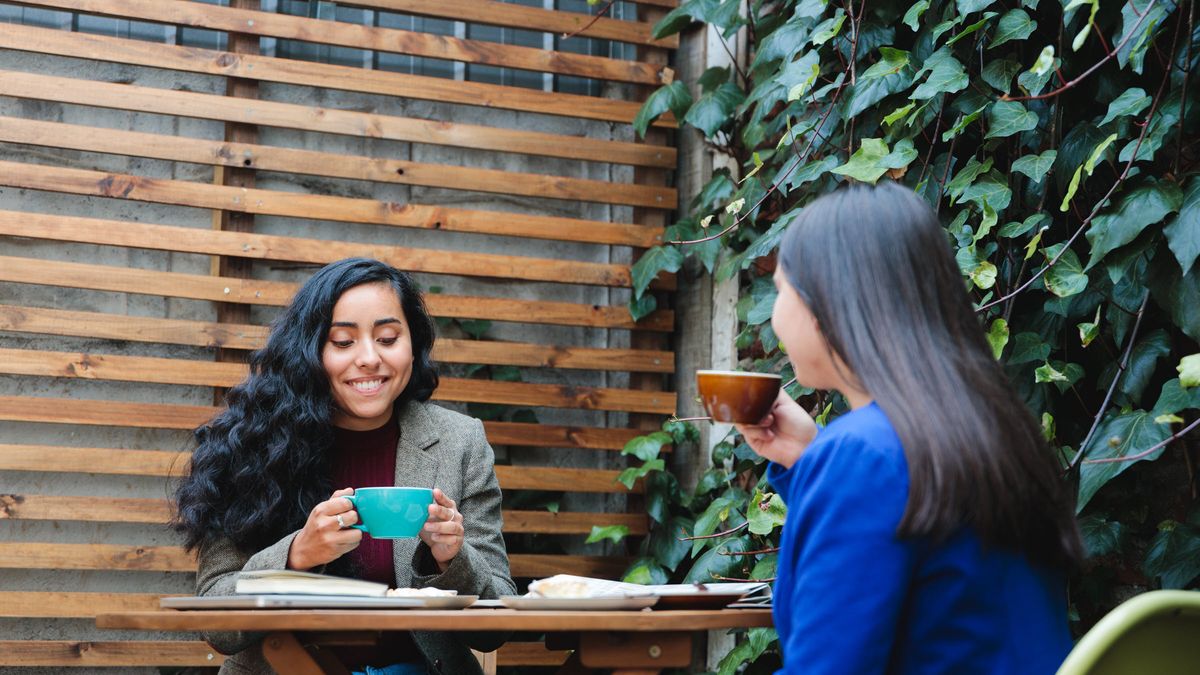 Cafe patio with lush foliage hosts two women enjoying coffee and pastries.
