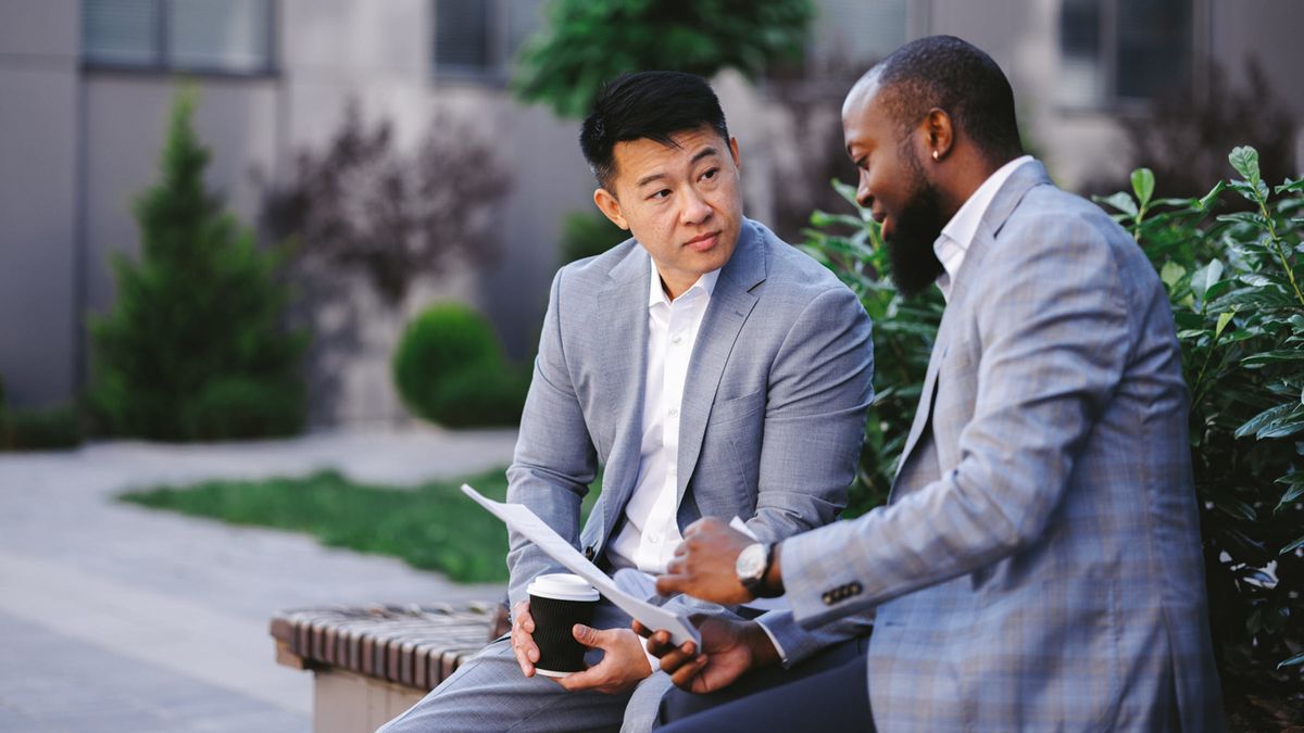 Side view of two colleagues outside office, reading information on a document and drinking coffee