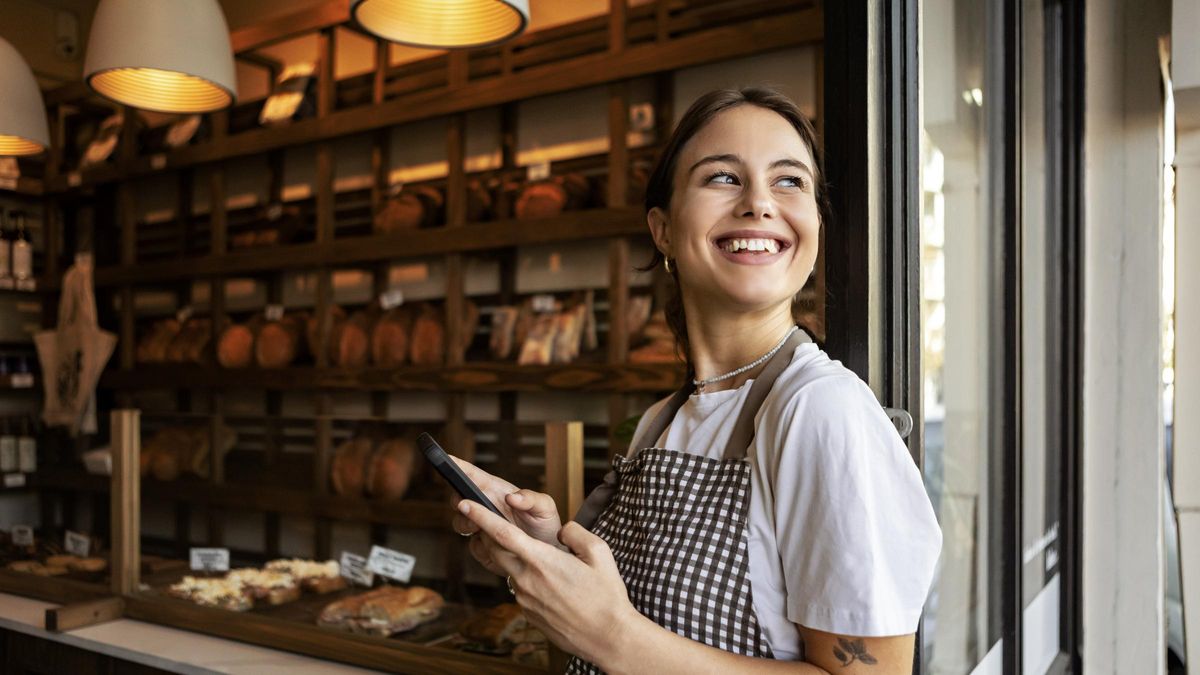 Young bakery owner standing proudly outside her shop using phone