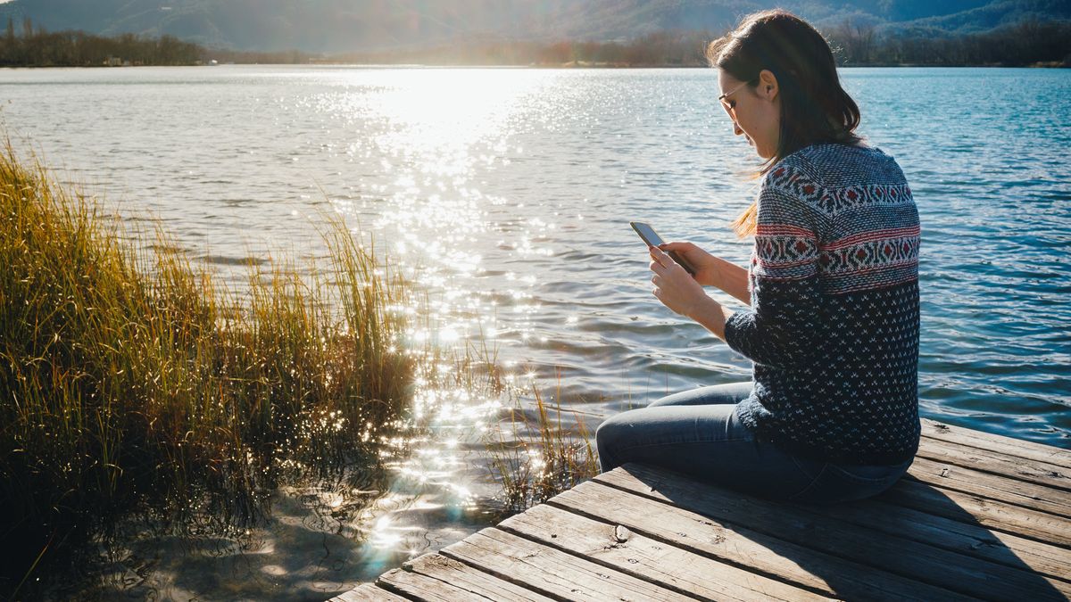 Woman sitting on a wooden platform at a lake at sunset, using smartphone
